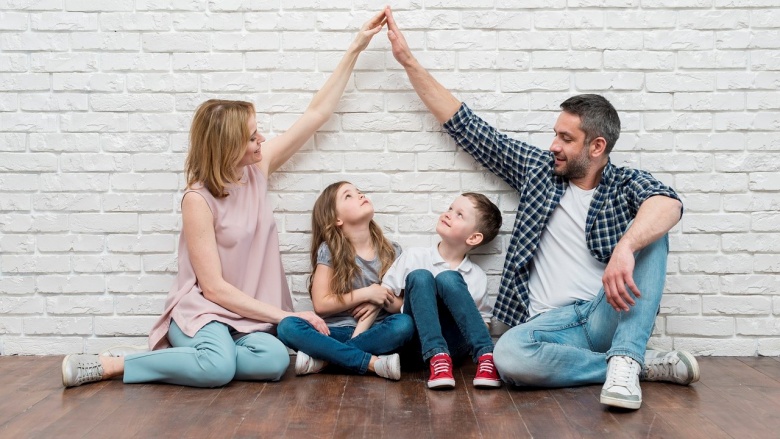 Vater und Mutter sitzen mit ihren beiden Kindern auf dem Fußboden und formen mit ihren Armen zusammen ein Dach.